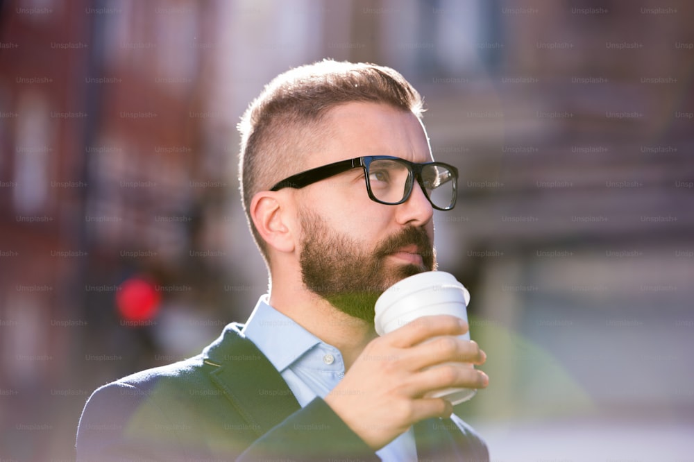 Uomo d'affari hipster che tiene una tazza usa e getta e beve caffè, cammina per le strade di Londra, indossando occhiali neri