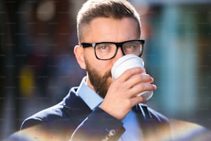 Uomo d'affari hipster che tiene una tazza usa e getta e beve caffè, cammina per le strade di Londra, indossando occhiali neri