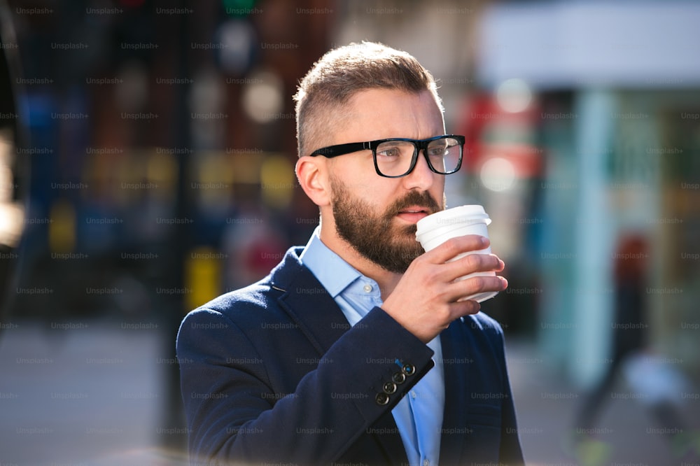 Hipster-Geschäftsmann, der einen Einwegbecher hält und Kaffee trinkt, durch die Straßen von London geht und eine schwarze Brille trägt