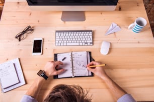 Businessman at the desk, wearing smart watch, writing into personal organizer.  Computer, smart phone and various office supplies around the workplace.