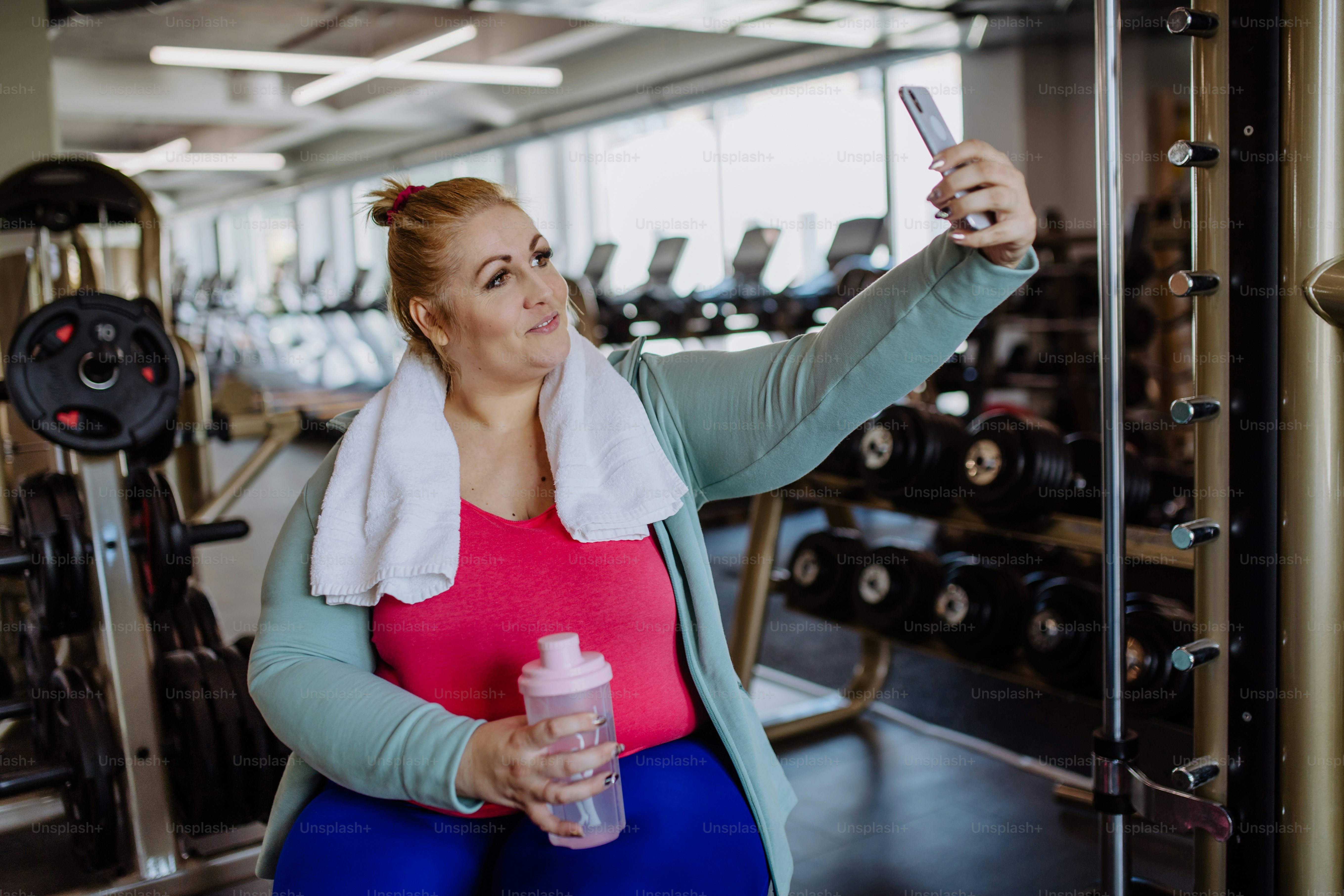 A happy mid adult overweight woman taking selfie after exercise indoors in gym
