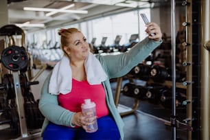 A happy mid adult overweight woman taking selfie after exercise indoors in gym