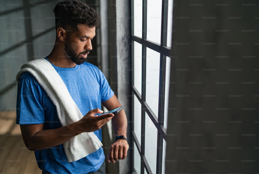 Ein junger afroamerikanischer Sportler, der drinnen im Fitnessstudio steht und Smartphone und Smartwatch benutzt.