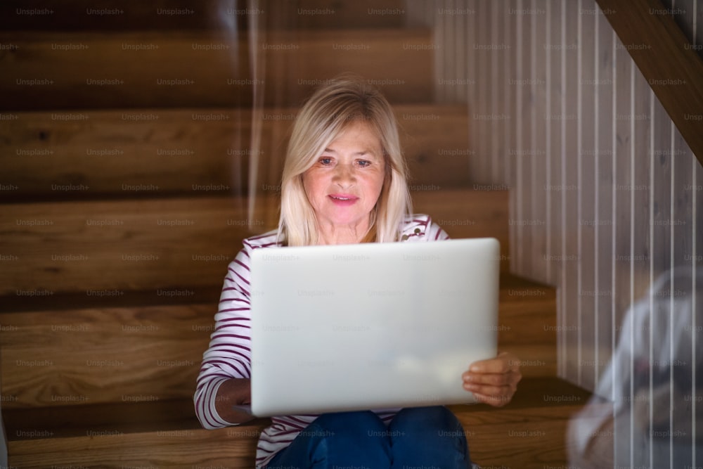 Vorderansicht einer älteren Frau mit Laptop, die drinnen zu Hause auf einer Treppe sitzt und arbeitet.