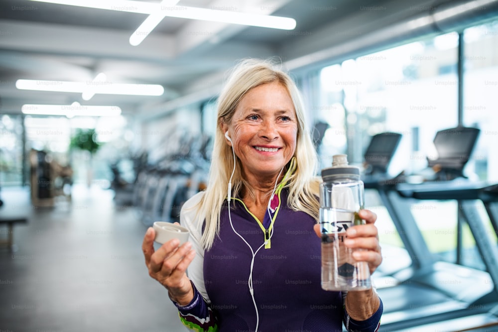 A front view of senior woman with earphones and water bottle in gym, resting after doing exercise.