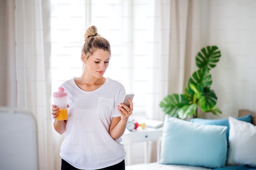 A young woman doing exercise indoors at home, using smartphone.