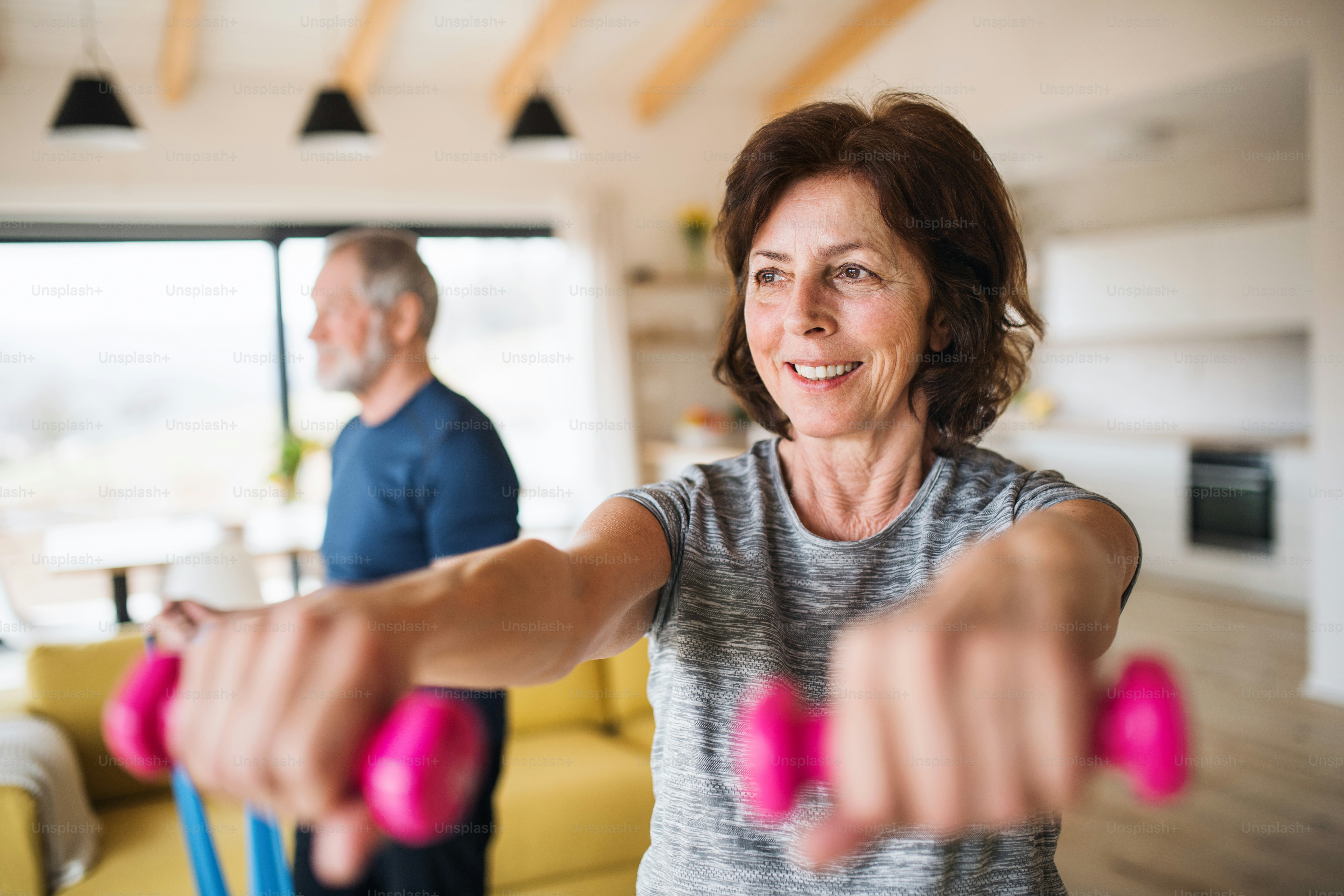A happy senior couple with dumbbells indoors at home, doing exercise indoors