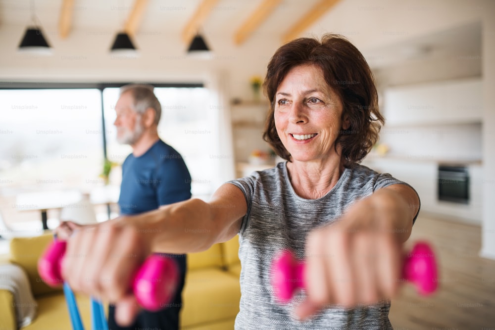 A happy senior couple with dumbbells indoors at home, doing exercise indoors.