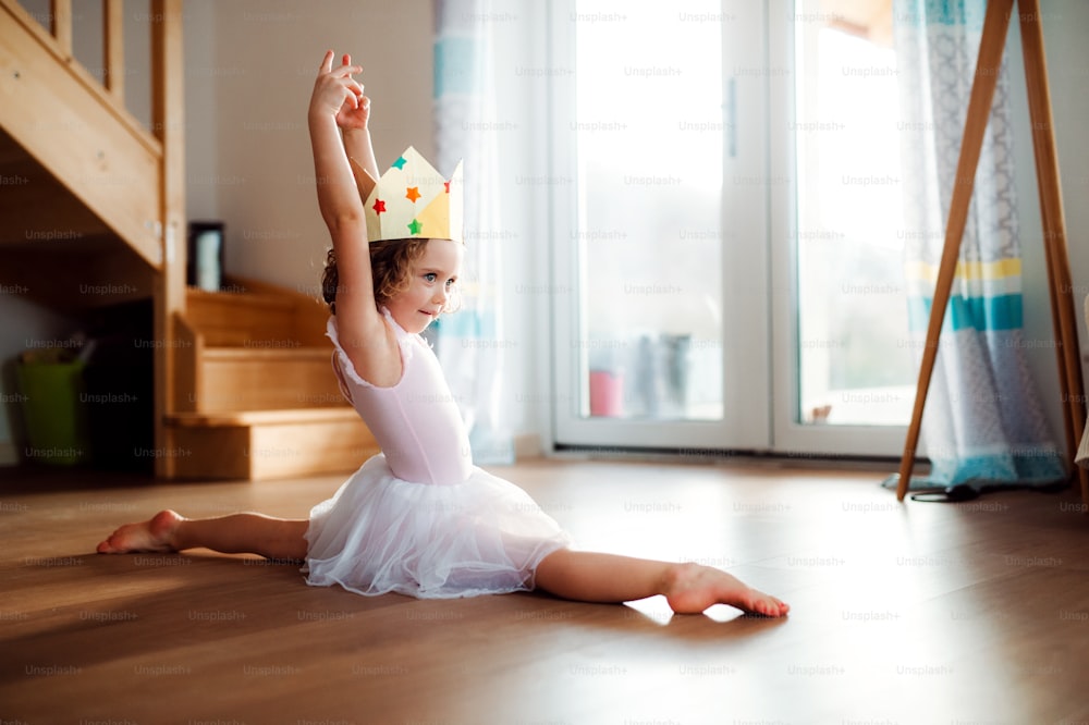 Una pequeña bailarina con una corona de princesa en casa, haciendo gimnasia dividida.