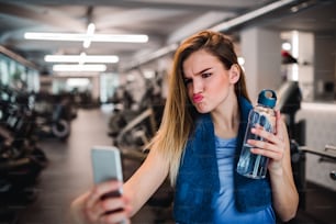 A portrait of young girl or woman with water bottle and smartphone in a gym, pouting lips and taking selfie.