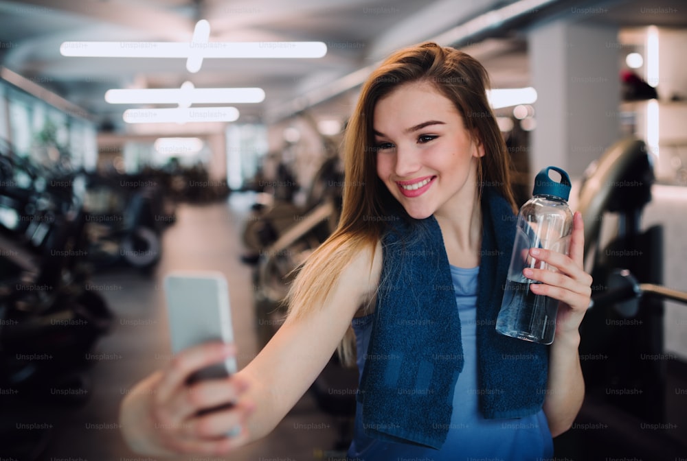 Un ritratto di ragazza o donna con bottiglia d'acqua e smartphone in una palestra, che scatta selfie.