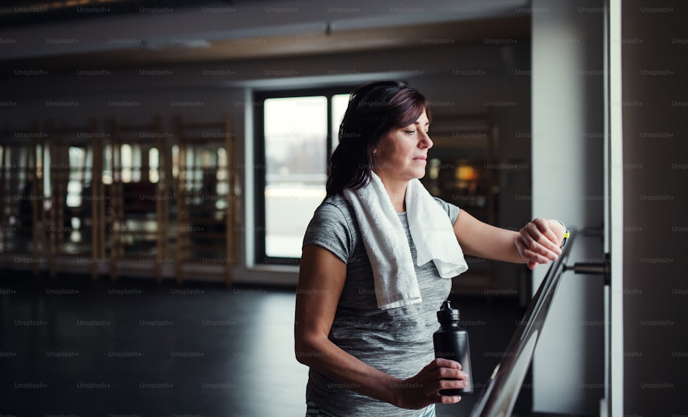 A content senior woman with water bottle in gym resting after doing exercise, checking the time. Copy space.