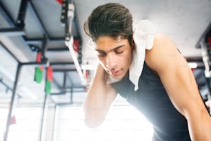 Young fit hispanic man in black sleeveless shirt in modern gym gym towel around his neck, wiping sweat off face