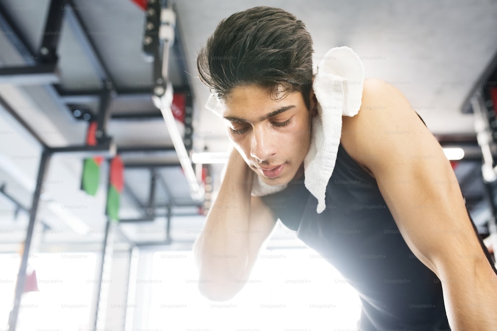 Young fit hispanic man in black sleeveless shirt in modern gym gym towel around his neck, wiping sweat off face