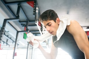 Joven hispano en forma con camisa negra sin mangas en una toalla moderna de gimnasio alrededor de su cuello, secándose el sudor de la cara