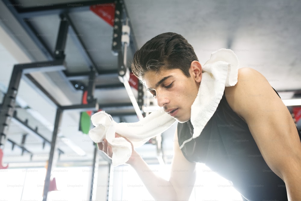 Jovem homem hispânico em forma de camisa preta sem mangas na toalha de ginástica moderna em torno de seu pescoço, limpando o suor do rosto