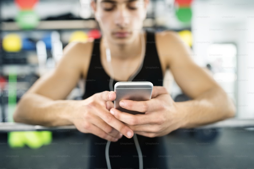 Hispanic fitness man in gym resting, holding smart phone, earphones in his ears listening music