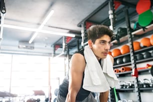 Jovem homem hispânico em forma de camisa preta sem mangas, na academia moderna, toalha em volta do pescoço, descansando
