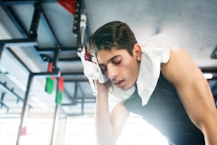 Joven hispano en forma con camisa negra sin mangas, en gimnasio moderno, toalla alrededor del cuello, secándose el sudor de la cara