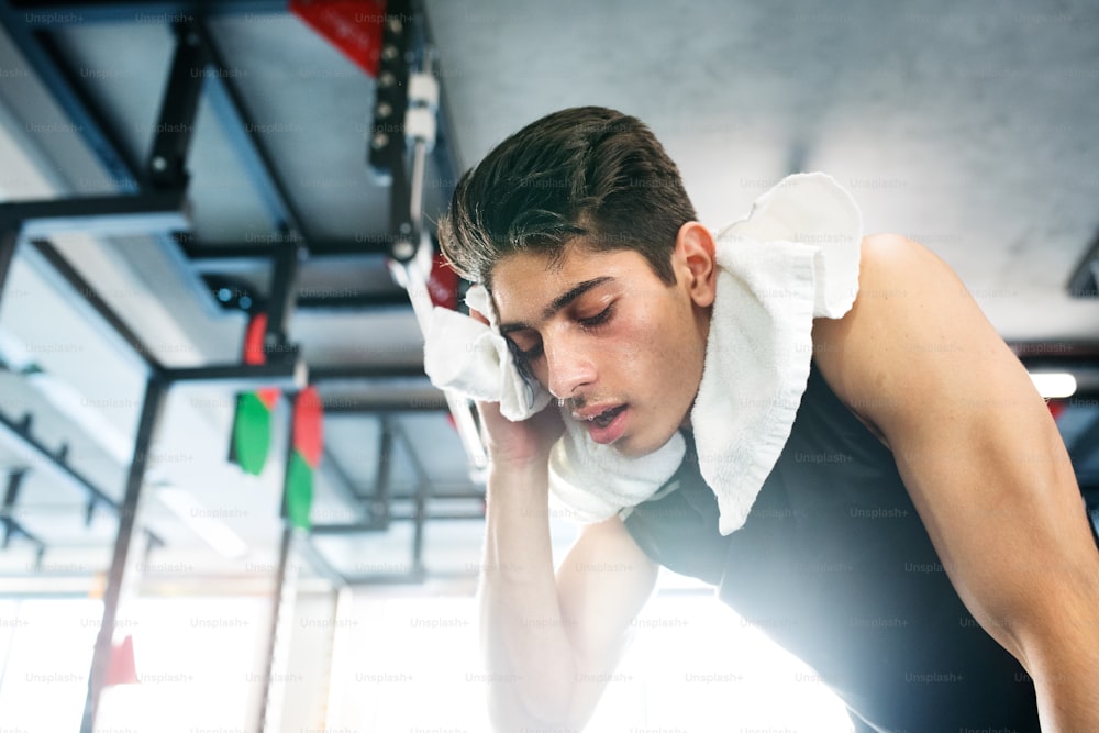 Young fit hispanic man in black sleeveless shirt, in modern gym gym, towel around his neck, wiping sweat off face