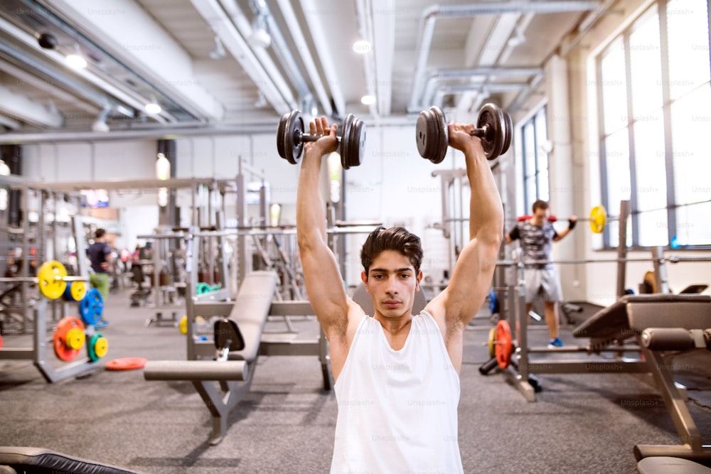 Jeune homme de fitness hispanique au gymnase assis sur un banc, s’entraînant avec des poids