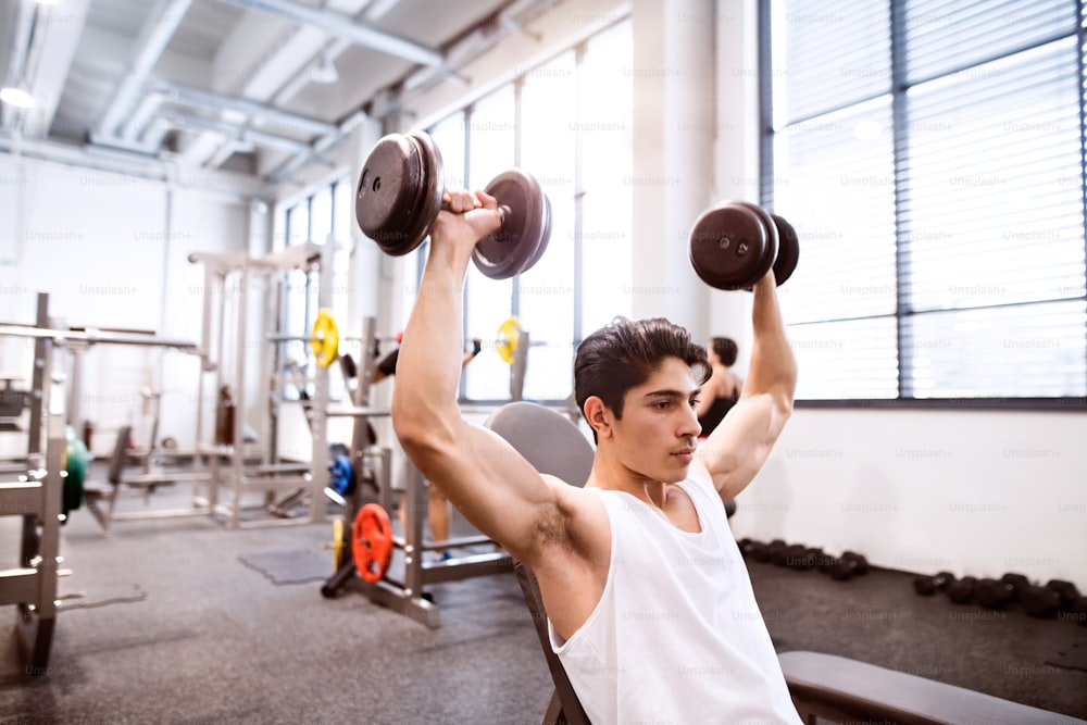 Junger hispanischer Fitnessmann im Fitnessstudio, der auf einer Bank sitzt und mit Gewichten trainiert