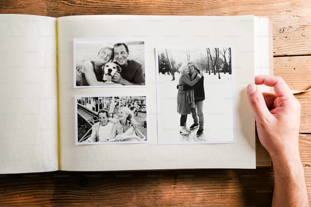 Hand of unrecognizable person holding a photo album looking at various black and white pictures of senior couple. Studio shot on wooden background.