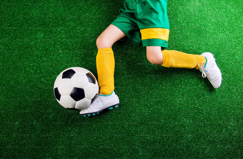Jambes d’un petit joueur de football méconnaissable avec un ballon de football contre du gazon artificiel. Photo en studio sur herbe verte.