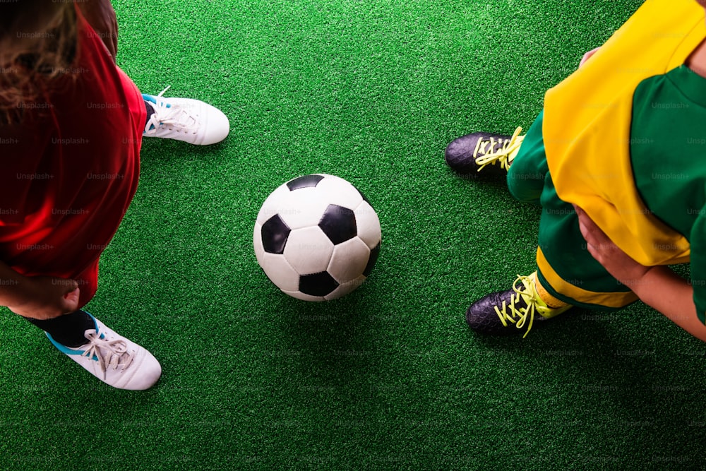 Legs of two unrecognizable little football players with soccer ball against artificial grass. Studio shot on green background.