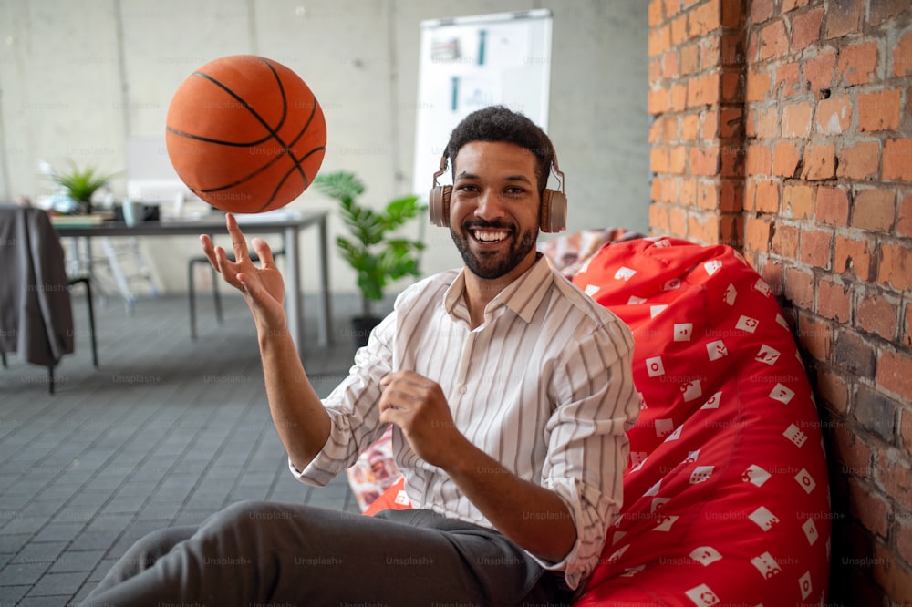 A cheerful young businessman with headphones and ball taking a break in office, resting and looking at camera.