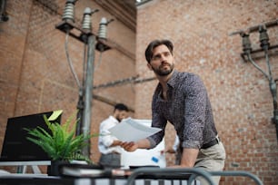 A thoughtful young businessman standing at desk in office, working.