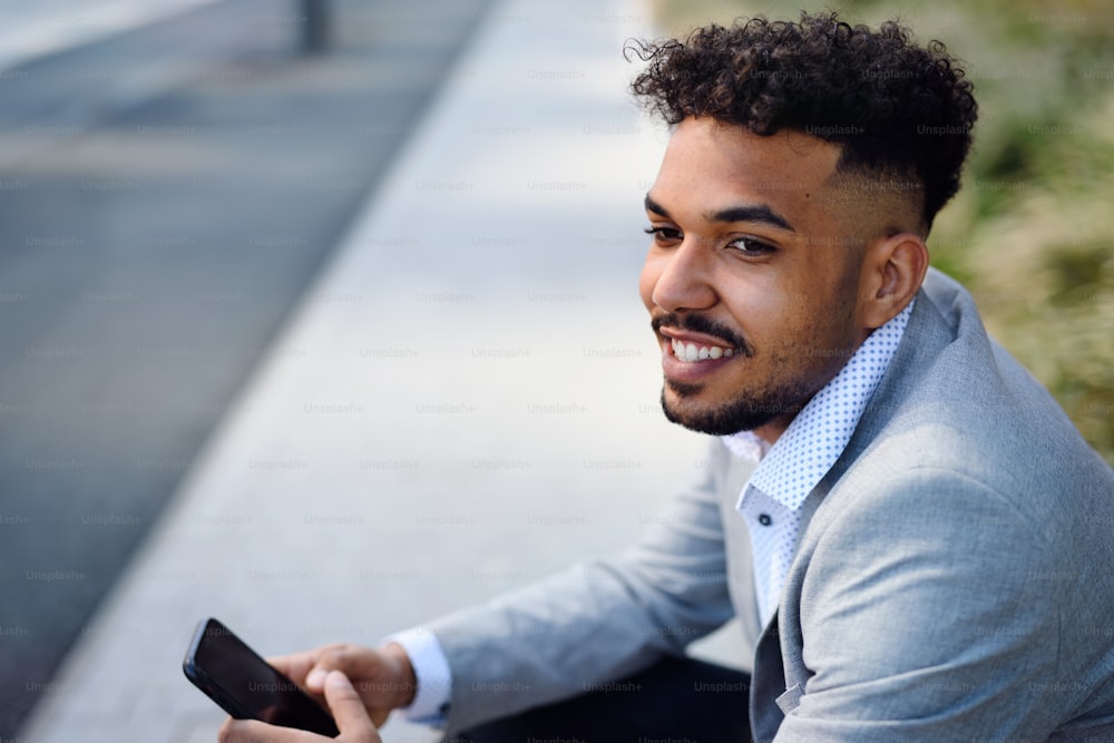 Ein Porträt eines jungen Mannes, der draußen in der Stadt sitzt und ein Smartphone benutzt.