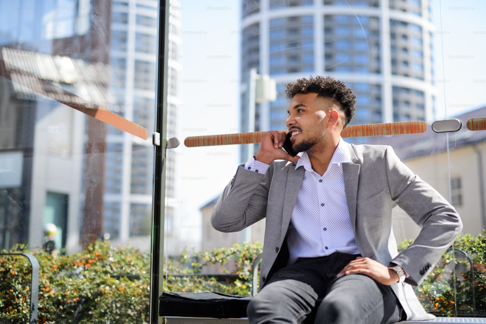 A portrait of young man student sitting outdoors on bus stop in city, using smartphone.