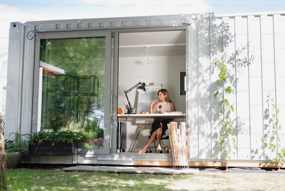 Front view of mature woman working indoors in home office in container house in backyard.