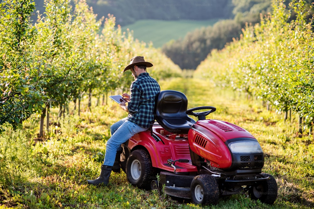 A mature farmer standing outdoors by mini tractor in orchard, using tablet.