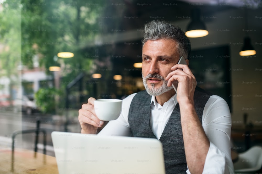 Un hombre maduro con una computadora portátil en la mesa en un café, usando un teléfono inteligente.