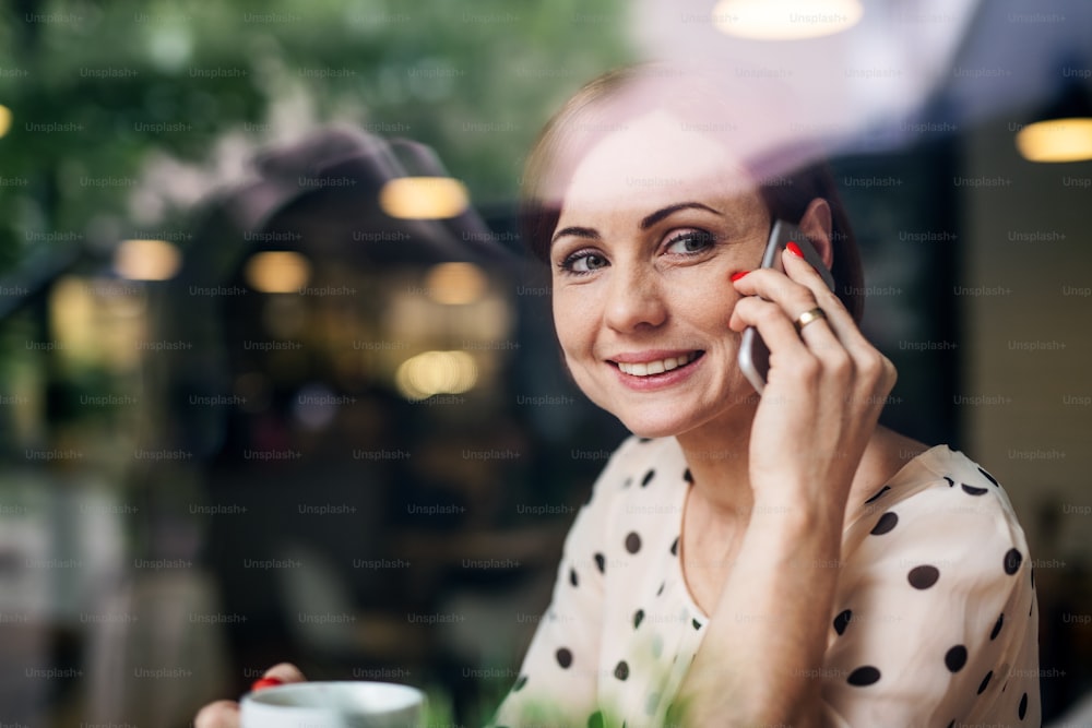 Un retrato de una mujer con café y teléfono inteligente sentada a la mesa en un café, haciendo una llamada telefónica. Disparado a través del vidrio.