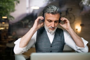 Mature man with headphones at the table in a cafe, using laptop. Shot through glass.