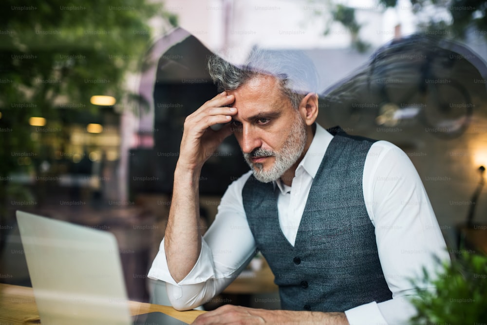 Serious mature man sitting at the table in a cafe, using laptop. Shot through glass.