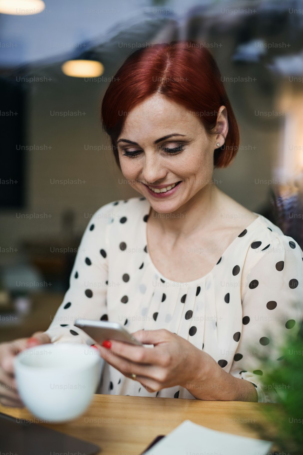 A portrait of woman with coffee and smartphone sitting at the table in a cafe, using laptop. Shot through glass.