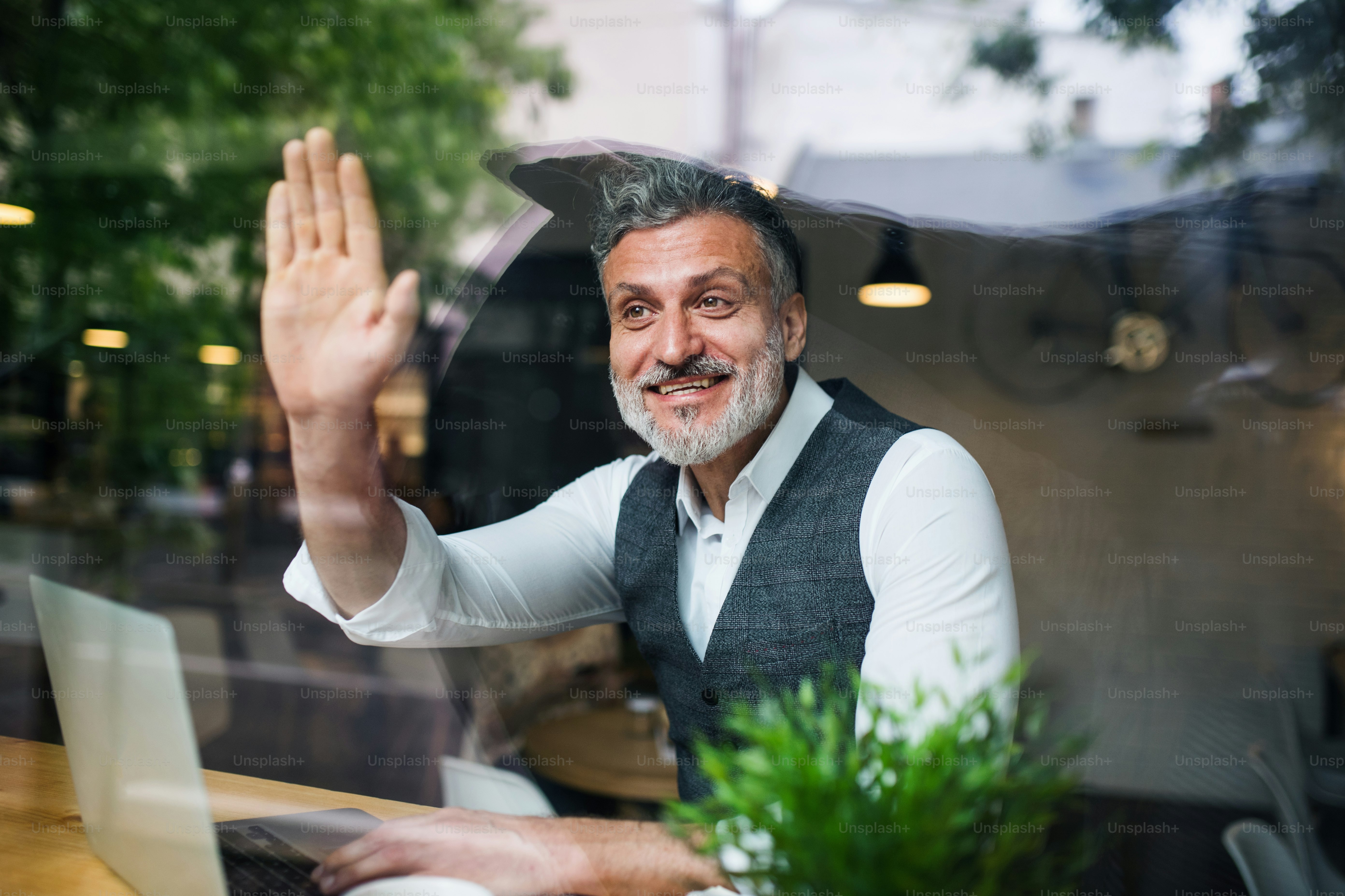 Mature man with laptop at the table in a cafe, greeting somebody. Shot through glass.