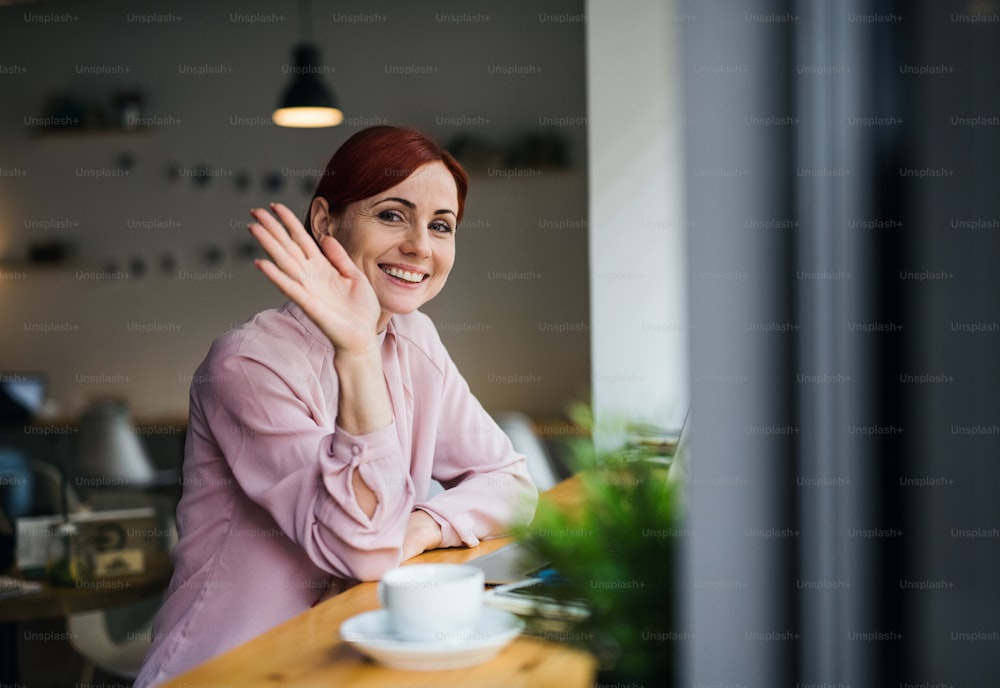 A portrait of attractive woman with coffee sitting at the table in a cafe, waving at somebody.