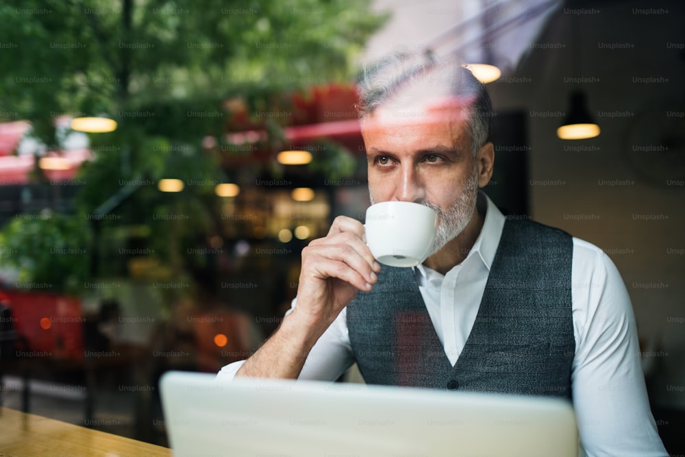 Reifer Mann mit Kaffee sitzt am Tisch in einem Café und benutzt einen Laptop. Durch Glas geschossen.