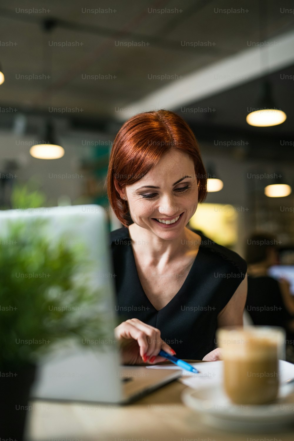 Ein Porträt einer attraktiven Frau mit Kaffee, die mit Laptop am Tisch in einem Café sitzt. Durch Glas geschossen.
