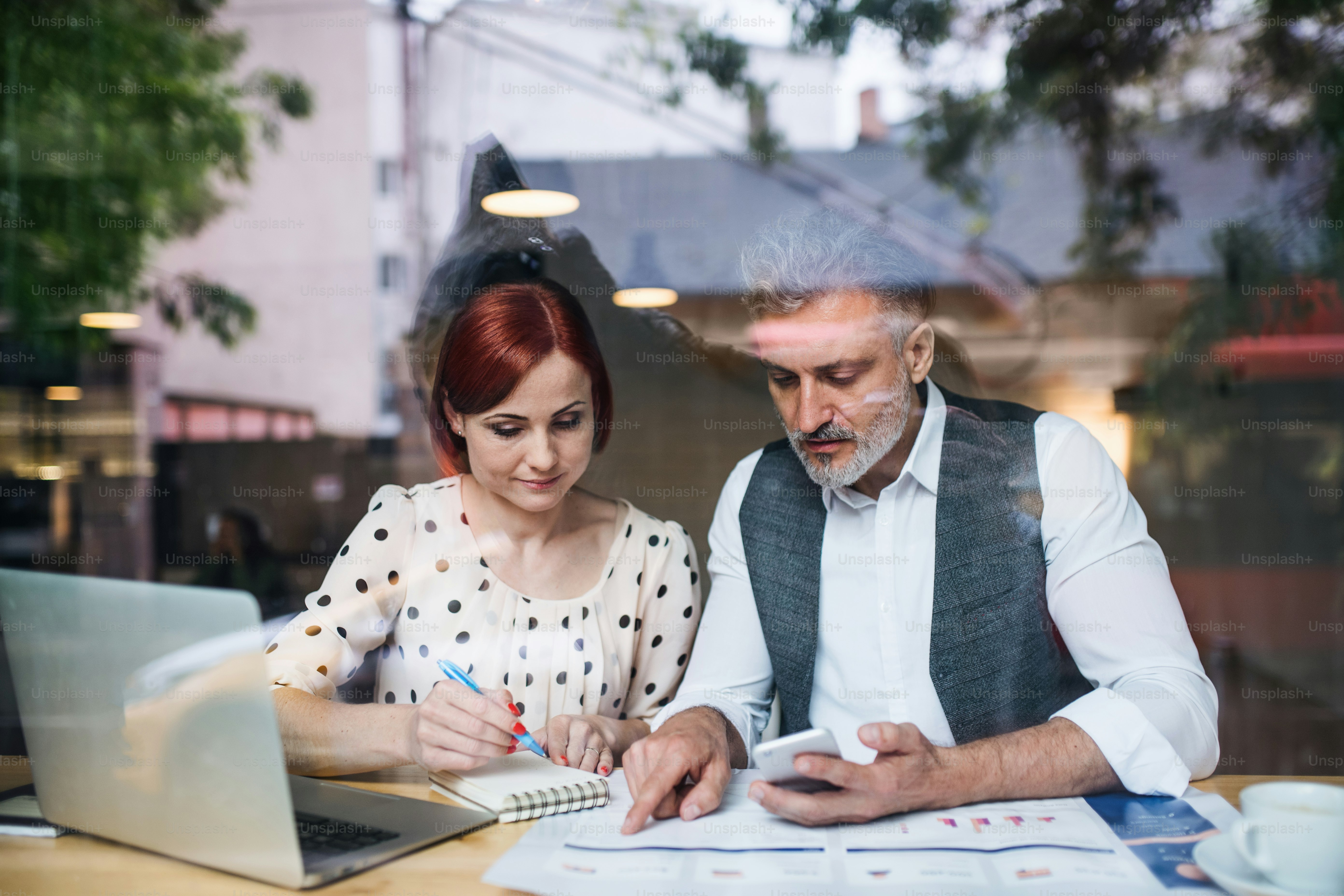 A man and woman having business meeting in a cafe, using laptop and smartphone. Shot through glass.