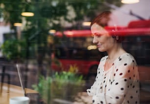 Un portrait d’une femme avec des écouteurs assise à la table d’un café, utilisant un ordinateur portable. Tiré à travers le verre.