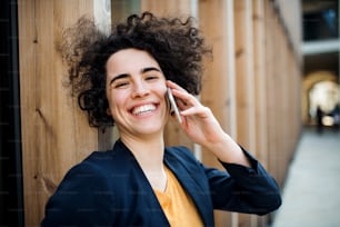 A happy young business woman with smartphone standing outdoors, making a phone call. Start-up concept.