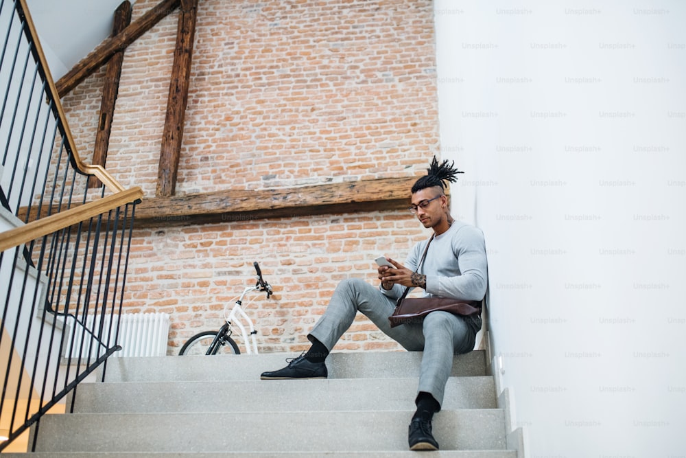 Un joven hombre de negocios de raza mixta con teléfono sentado en las escaleras de un edificio de oficinas.