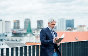 A mature businessman with diary standing on a terrace outside office, writing and planning.