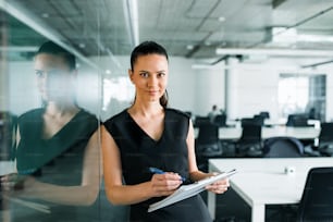 Una vista frontal de una joven empresaria con portapapeles en una oficina, de pie.